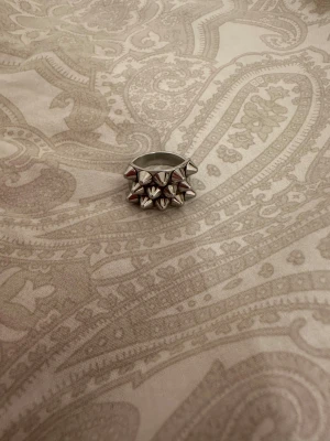 Silvrig ring med nitar. PRIS KAN DISKUTERAS - Cool och chunky ring i silverfärgad metall med flera små konformade nitar på ovansidan. Ringen har en bred och robust form som ger en edgy vibe, perfekt för att lyfta din stil. Passar dig som gillar att sticka ut och addera lite attityd till outfiten.