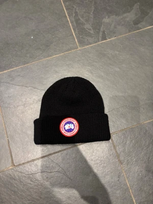 Svart stickad mössa från Canada Goose - Svart ribbstickad mössa från Canada Goose med klassisk rund logotyp-patch framtill. Mössan har uppvikt kant och är tillverkad i mjukt stickat material som håller dig varm. Perfekt för kalla dagar och snygg till streetwear.