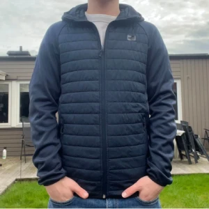 Mörkblå quiltad pufferjacka med huva - Snygg mörkblå pufferjacka från Jack & Jones. Jackan har två sidofickor med dragkedja. Perfekt för dig som gillar sportig och clean stil.