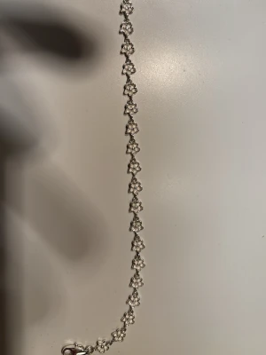 Silverfärgat armband med blommor från MULI - Snyggt armband i silverfärg med små blommor längs hela kedjan. Blommorna är detaljerade och ger ett fräscht och stilrent intryck. Armbandet har en klassisk karbinhake och en liten rund bricka vid låset. Perfekt för dig som gillar blommiga accessoarer.