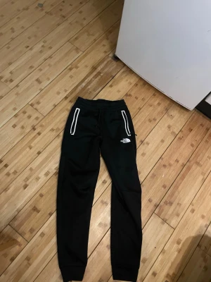 Svarta mjukisbyxor från The North Face - Svarta mjukisbyxor från The North Face med vita detaljer runt fickorna och logga på benet. Byxorna har snörning i midjan och ribbade muddar vid bensluten. Perfekta för chill eller träning, riktigt sköna och snygga.