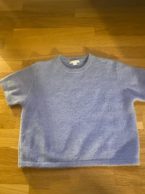 Ljusblå stickad topp från Gina Tricot - Supermysig ljusblå stickad topp från Gina Tricot med korta ärmar och rund hals. Mjuk och fluffig känsla, perfekt för att ge din outfit en chill vibe. Passar dig som gillar enkel och stilren look med lite cozy touch.