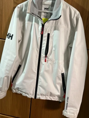 Vit vindjacka från Helly Hansen M - Snygg vit vindjacka från Helly Hansen med svarta detaljer och röda dragkedjor. Jackan har hög krage, flera fickor med dragkedja och logga på ärm och krage. Materialet är lätt och syntetiskt, perfekt för blåsiga dagar. Stilren design med normal passform och lång ärm.