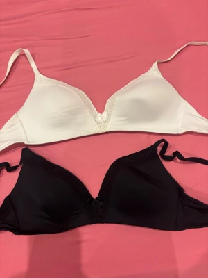 Svart och vit spetsdetaljerad bh - Två snygga bh, en i svart och en i vit, båda med spetsdetaljer längs v-ringningen och en liten rosett framtill. Bhn har släta kupor och tunna axelband, perfekta för att ge en clean och chic look.