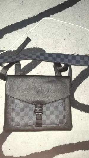 Svart axelväska rutig - Snygg svart axelväska  med klassiskt rutigt mönster och justerbar axelrem. Väskan är tillverkad i skinn och har ett lock med spänne framtill. Perfekt för dig som vill ha en stilren och trendig accessoar.