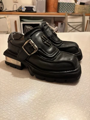 Svarta New Rock platåskor i skinn - Säljer ett par chunky New Rock platform skor i svart skinn med grov sula och metallklack. Skorna har en bred rem med metallspänne och New Rock-logga på plösen. Fyrkantig tå och metal detaljer