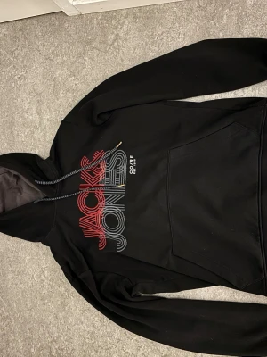 Jack & Jones Hoodie  - Jack & Jones hoodie Stilren svart hoodie med tydlig Jack & Jones-logga framtill – Jack i röd text och Jones i grå. Tillverkad i mjukt och bekvämt material med skön passform. Enkel att matcha och perfekt för både vardag och fritid. En tidlös hoodie i bra skick. M men funkar som S