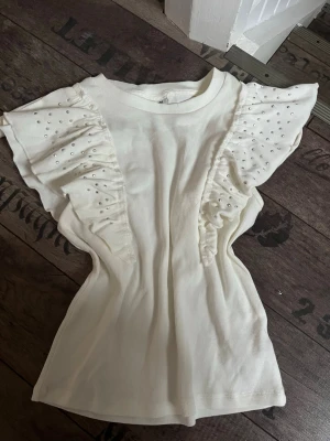 Vit topp med volangärmar och pärlor - Säljer en vit topp med korta volangärmar dekorerade med små pärlor. Toppen har rund hals och är tillverkad i mjukt ribbat material som ger en skön känsla. Perfekt för dig som gillar detaljer och vill ha en söt och trendig look.