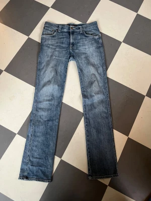 Blå Nudie Jeans  - Snygga blå jeans från Nudie Jeans med klassisk femficksdesign och raka ben. Jeansen har en cool tvättad look, orangea detaljer på bakfickorna och logopatch i läder baktill. Passar dig som gillar tidlös stil och sköna material. Modell slim Jim storlek W29 L32