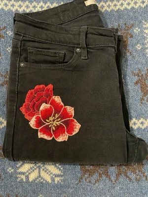 Svarta bootcut jeans med broderade blommor - Svarta jeans med bootcut passform och snygga slitningar vid knäna. På framsidan och bakfickan finns broderade röda blommor som ger en unik vibe. Jeansen har klassisk femficksdesign och är gjorda i jeansmaterial med stretch.