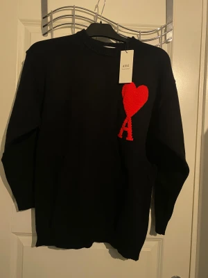 Ami Paris Sweater HELT NY!!! - Svart stickad tröja från Ami Alexandre Mattiussi med rund hals och lång ärm. På bröstet finns ett stort rött hjärta med bokstaven A, vilket ger en unik och cool look. Tröjan har ribbade muddar vid hals, ärmslut och nederkant.