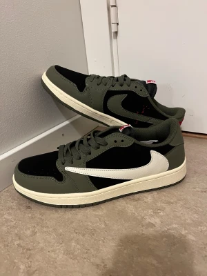 Nike Air Jordan 1 Low Travis Scott Olive - Nike Air Jordan 1 Low Travis Scott i olivgrönt och svart med vit swoosh och röda broderade detaljer på hälen. Snygg kontrast mellan färgerna och klassisk Jordan-vibe. Skorna har snörning, platt sula och rund tå. Materialet är skinn och syntet.