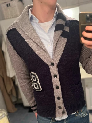 Replay stickad kofta  - Snygg stickad kofta från Replay i mörkblått och grått med bred krage och knappar framtill. Har coolt broderat nummer 8 på ena sidan och två fickor. Randiga detaljer på kragen och ärmslut ger extra stilpoäng. Perfekt för dig som gillar retro college-vibe. Den är lite nopprig