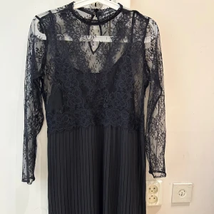 Ny Svart spetsklänning från Zara - Supersnygg svart klänning från Zara med transparent spets upptill och långa ärmar. Överdelen har blommiga spetsdetaljer och knäppning i nacken, medan kjolen är plisserad för en extra cool look. Perfekt för dig som vill sticka ut med stil. Klänningen är som S i storlek 