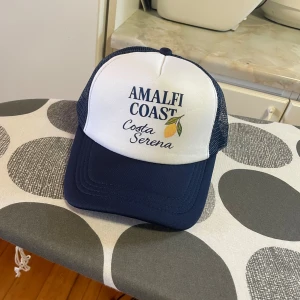 Amalfi Coast truckerkeps blå/vit - Säljer en snygg truckerkeps med texten 'AMALFI COAST Costa Serena' och citronmotiv. Kepsen har vit frontpanel och mörkblått nät på sidor och bak, samt böjd skärm i mörkblått. Perfekt för sommarens äventyr och chill dagar.