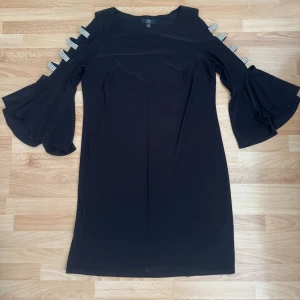 Svart klänning med glittriga detaljer - Svart klänning från JBS  med glittriga band längs ärmarna och snygga volangärmar. Klänningen har rund halsringning och är tillverkad i mjukt material. Perfekt för dig som gillar att sticka ut med coola detaljer och glam. Storleklapp avklippt, men den är storlek M, bröst 98-100cm. Använd en gång.