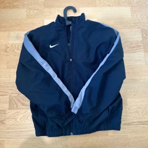 Svart Nike vindjacka med grå detaljer - Sportig svart vindjacka från Nike med grå ränder längs ärmarna och klassisk vit swoosh på bröstet. Jackan har hel dragkedja, hög krage och resår vid ärmslut och midja. Perfekt för en aktiv stil och enkel att matcha.