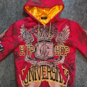Röd Hoodie från Christian Audigier - Tuff röd hoodie från Christian Audigier med stort Hip Hop University-tryck, vingar och krona på bröstet. Guldfärgad snörning, dragkedja med metallberlock och guldfärgat foder i huvan. Långa ärmar med coola detaljer och färgglada motiv på ärmarna.