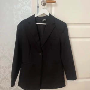 Svart kavaj från H&M Divided XXS - Klassisk svart kavaj från H&M Divided i storlek XXS. Enkelknäppt med en knapp framtill, smala slag och tre fickor. Snygg och stilren modell som passar perfekt till kostymbyxor eller jeans. Materialet är mjukt och följsamt.