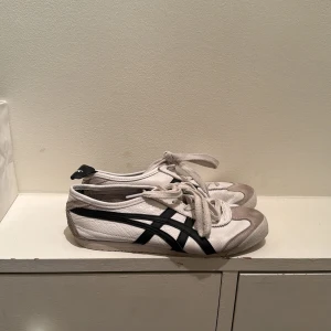 Onitsuka Tiger Mexico 66 vita sneakers - Säljer ett par klassiska Onitsuka Tiger Mexico 66 sneakers i vitt skinn med svarta detaljer och grå mocka vid tån. Skorna har snörning, platt sula och den ikoniska svarta randen på sidorna. Perfekta för dig som gillar retrostil och streetwear. Ena lappen bak saknas.