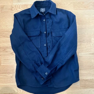 Mörkblå overshirt från H&M - Stilren mörkblå overshirt från H&M med klassisk krage, två stora bröstfickor och knäppning framtill. Skjortan har lång ärm och är tillverkad i ett mjukt bomullstyg. Perfekt att bära öppen eller stängd för en avslappnad look.