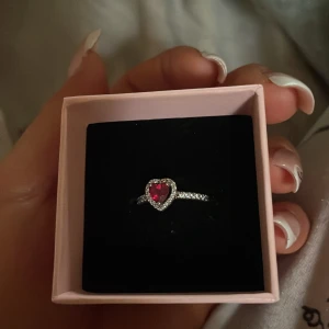 Pandora silverring med hjärta - Superfin silverring från Pandora med ett rött hjärta i mitten, omgiven av små glittrande stenar. Ringen har en elegant och romantisk design som verkligen sticker ut. Perfekt för dig som älskar detaljer och vill ha något extra speciellt.  storlek 52!  som ny knappas använd.
