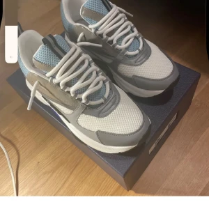 Ljusblåa och vita B22 dior - Säljer ett par snygga sneakers i grått, vitt och blått med coola detaljer. Ovandelen är i mesh och syntet, med vita snören och en chunky vit sula med svarta och blå inslag. Skorna har rund tå och sportig vibe, perfekta för dig som gillar streetstyle.