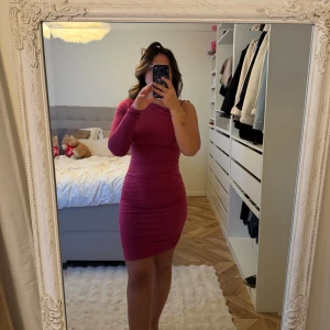 Mörkrosa one shoulder kortklänning - Säljer en tight mörkrosa kortklänning med one shoulder-design och lång ärm på ena sidan. Klänningen har en figurnära passform och är tillverkad i ett stretchigt material som framhäver siluetten. Perfekt för dig som vill sticka ut med en unik look.