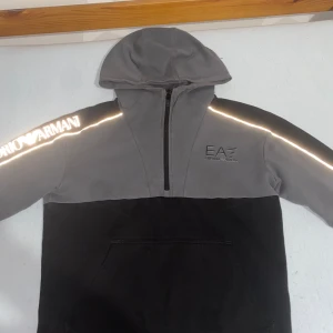 Grå och svart EA7 hoodie med huva - Säljer en snygg tvåfärgad hoodie från EA7 Emporio Armani. Tröjan är grå upptill och svart nedtill, har huva, halv dragkedja och reflekterande logga på ärmen. Perfekt för dig som gillar streetwear och vill sticka ut med stilren design. 