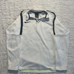 Vit Nike Dri-Fit långärmad tröja - Vit långärmad Nike Dri-Fit tröja med svarta detaljer längs axlarna och dragkedja vid halsen. Perfekt för träning eller sportiga dagar. Materialet är lätt och andas, vilket gör den skön att ha på sig under aktivitet.