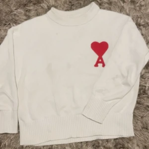 Vit stickad tröja från Ami Paris - Vit stickad tröja från Ami Paris med röd hjärtlogga och bokstaven A på bröstet. Tröjan har ribbade muddar och en klassisk rund halsringning. Perfekt för dig som gillar stilrena plagg med en twist.