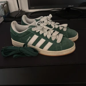 Adidas Campus gröna sneakers mocka - Säljer ett par Adidas Campus sneakers i mörkgrön mocka med klassiska vita ränder och vit snörning. Skorna har en platt gummisula i beige och kommer med extra gröna skosnören. Rund tå och låg profil, perfekt för dig som gillar retrostil. Skolådan ingår också