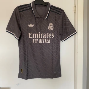 Real Madrid x Adidas fotbollströja M - Snygg mörkgrå Real Madrid fotbollströja från Adidas med klassisk krage och knappar. Tröjan har broderat klubbmärke, Adidas-logga och tryckta detaljer i vitt. Tillverkad i lätt och ventilerande material, perfekt för match eller träning.