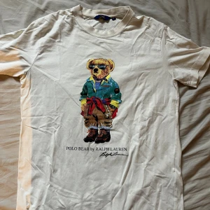 Vit Polo Bear t-shirt från Ralph Lauren - Vit t-shirt från Ralph Lauren med ikoniskt Polo Bear-tryck framtill. Björnen har coola solglasögon och färgglada kläder. 