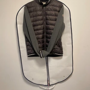 Grå Moncler cardigan  med dragkedja - Snygg cardigan  från Moncler i mörkgrått med quiltad front och ärmar i mjukare grått tyg. Jackan har hel dragkedja, ribbade muddar med vit och svart rand samt klassisk Moncler-logga på ärmen. Perfekt för dig som vill ha en stilren och sportig look. Den sitter som en M är 182 och den sitter fint 