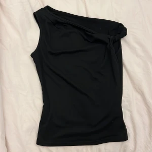 Svart off shoulder topp med knut - Säljer en svart one shoulder topp med snygg knutdetalj på axeln. Toppen är ärmlös och har en tight passform som framhäver siluetten. 