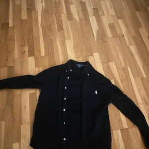 Mörkblå skjorta från Ralph Lauren - Stilren mörkblå skjorta från Ralph Lauren med klassisk krage och vita knappar. Skjortan har lång ärm och den ikoniska broderade loggan på bröstet. Perfekt för dig som gillar en clean och tidlös look.