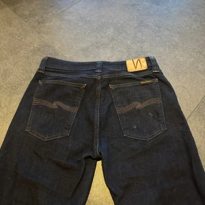 Mörkblå jeans från Nudie Jeans, 31/32 - Säljer ett par mörkblå jeans från Nudie Jeans i klassisk rak modell. Jeansen har snygga bakfickor med Nudie-signatur och läderpatch i midjan. Tillverkade i jeansmaterial av bomull, passar dig som gillar stilrena och tidlösa jeans.