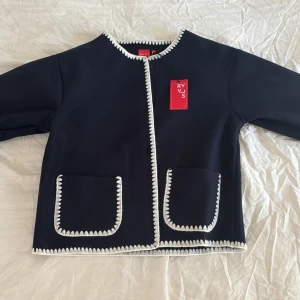 Mörkblå cardigan jacka från RYVLS - Mörkblå cardigan jacka från RYVLS med vita virkade kanter längs ärmar, fickor och öppning. Jackan har två stora fickor framtill och är långärmad med en rak, boxig passform. Materialet är mjukt och känns som ull. Perfekt för dig som gillar stilrena detaljer.