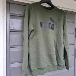 Olivgrön sweatshirt från Puma - Olivgrön sweatshirt från Puma med klassisk logga i svart framtill. Tröjan har rund hals, ribbade muddar och är tillverkad i mjuk bomullsblandning. Perfekt för en avslappnad och sportig stil.