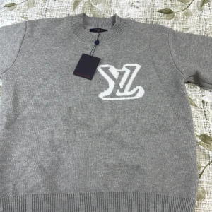 Grå stickad Louis Vuitton tröja - Snygg grå stickad tröja från Louis Vuitton med stort vitt LV-logo både fram och bak. Tröjan har rund halsringning och ribbade muddar vid ärmslut och nederkant. Perfekt för dig som vill ha en exklusiv och stilren look. 