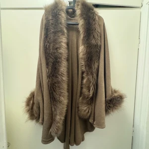 Beige kappa med fuskpälsdetaljer - Säljer en beige kappa/poncho med breda fuskpälsdetaljer längs krage och ärmslut. Jackan har en loose passform. Nypris - 1500kr
