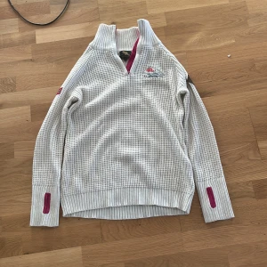 Vit stickad tröja Beaver Lake L - Vit stickad tröja från Beaver Lake med broderi 'Å i Lofoten' på bröstet. Tröjan har hög krage, halv dragkedja med rosa detaljer och ribbade muddar. Snyggt rutmönster i stickningen och långärmad modell. Perfekt för kyliga dagar.