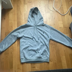 Grå hoodie från Ralph Lauren - Säljer en stilren grå hoodie från Ralph Lauren med klassisk huva och dragsnören. Tröjan har en broderad logga på bröstet, känguruficka och ribbade muddar. Perfekt för chill dagar och enkel att matcha med jeans eller joggers.