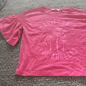 Rosa topp med puffärm och tryck - Säljer en rosa topp med puffiga korta ärmar och smockdetalj vid axlarna. Framsidan har ett tryck med texten 'California Surfing Paradise Los Angeles' och palmer. Toppen är i mjuk bomull och har en avslappnad passform.