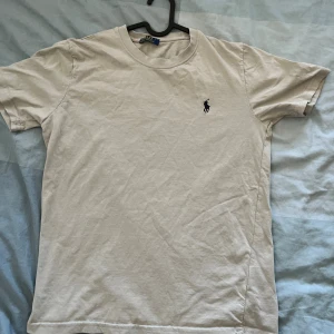 Beige t-shirt från Polo Ralph Lauren - Snygg beige t-shirt från Polo Ralph Lauren med klassisk rund hals och broderad marinblå logga på bröstet. Tillverkad i 100% bomull för en skön och mjuk känsla. Perfekt basic-plagg med stilren design.
