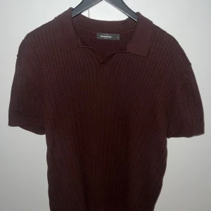 Brun stickad pikétröja Dressmann XL - Säljer en brun stickad pikétröja från Dressmann i storlek XL. Tröjan har korta ärmar, klassisk krage och ett snyggt kabelstickat mönster som ger en retrovibe. Perfekt för dig som gillar stilrena och bekväma plagg med lite extra struktur.