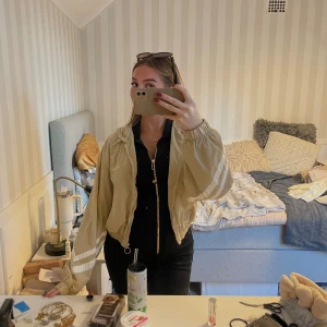 Beige vindjacka från Bershka - Säljer en beige vindjacka från Bershka med huva, dragsko och coola vita ränder på ärmarna. Jackan har croppad passform, dragkedja med ringdetalj och broderad text 'Extr.Street' på bröstet. Perfekt för vår eller höst och riktigt streetstyle-vibe.