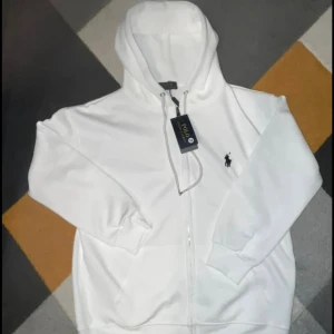 Vit hoodie från Polo Ralph Lauren - Snygg vit hoodie från Polo Ralph Lauren med dragkedja och klassisk logga broderad på bröstet. Tröjan har huva med snörning och två fickor framtill. Perfekt för en clean och stilren look.