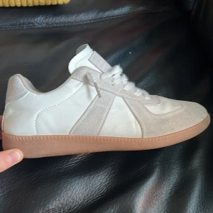 Vita sneakers med beige detaljer  - Snygga vita sneakers med beige mockadetaljer och ljusbrun gummisula. Skorna har rund tå, snörning och en stilren design som passar till många olika outfits. Materialet är en mix av skinn och mocka för en fräsch och modern look.